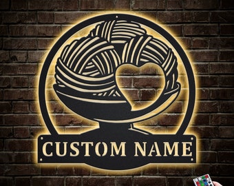 Pode incluir: Um letreiro de parede de metal preto com um design de novelo de lã e coração. O letreiro é iluminado com luzes LED branco quente e tem uma placa de nome personalizável que diz "CUSTOM NAME".