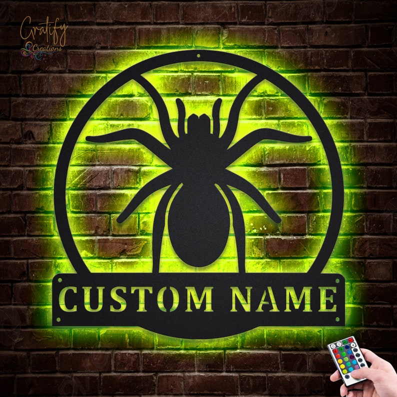 May include: A black metal spider silhouette with a custom nameplate, illuminated with a green light. The spider is in a circle with a web design.