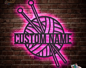 Pode incluir: Uma bola de lã e agulhas de tricô de metal preto com um brilho rosa, com o texto "CUSTOM NAME" no centro. A imagem está em um fundo de parede de tijolo.