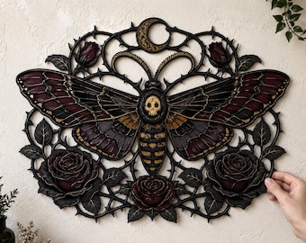 May include: A decorative wall hanging featuring a large moth with a skull pattern on its thorax, surrounded by dark red roses and thorny vines. A crescent moon sits at the top center. The piece is crafted in black with gold and red accents.