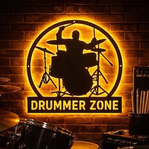 May include: A backlit metal sign featuring a drummer silhouette within a circle, with the words "DRUMMER ZONE" below. The sign is mounted on a brick wall, with drum sets and equipment visible in the foreground. The sign is illuminated with a warm, orange glow.