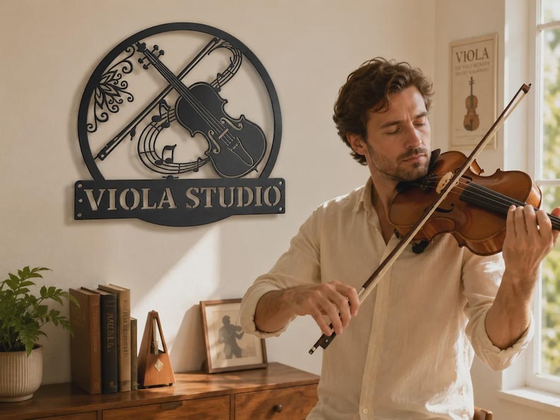 Puede incluir: Un letrero de pared de metal negro con un marco circular que presenta un dise&ntilde;o recortado de un viol&iacute;n, un arco y notas musicales, con el texto VIOLA STUDIO mostrado en una base rectangular en la parte inferior del marco.