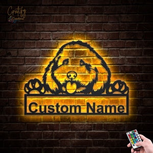 Puede incluir: Una silueta de perro de metal negro con sus patas sobre un letrero rectangular que dice "Custom Name". El letrero y el perro están iluminados con una luz amarilla cálida. Un control remoto con varios botones de colores es visible en la esquina inferior derecha de la imagen.