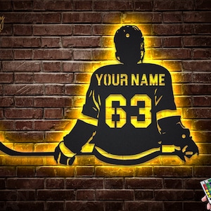 May include: A black silhouette of a hockey player holding a stick with a yellow glow around the figure. The player's jersey has the text "YOUR NAME" and the number "63". A remote control with multiple colored buttons is visible in the bottom right corner.
