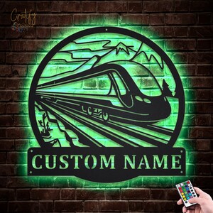 May include: A black metal silhouette of a train travelling through a mountain landscape with a green light shining through the metal. The silhouette is in a circle with the words "CUSTOM NAME" below it.