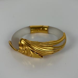 May include: A gold-coloured bracelet with a clear, circular band. The bracelet features a decorative gold leaf design on the front. The clasp and accents are also gold. The jewellery is set against a white background.
