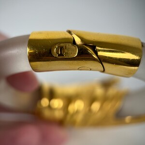 May include: Close-up of a gold-toned bracelet clasp with a clear, frosted band. The clasp features a small, embossed design and a secure closure. The bracelet is a fashion accessory.