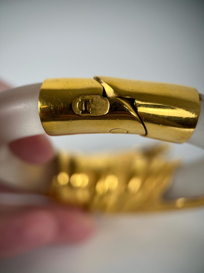 May include: Close-up of a gold-toned bracelet clasp with a clear, frosted band. The clasp features a small, embossed design and a secure closure. The bracelet is a fashion accessory.