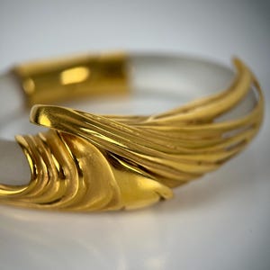 May include: A clear, transparent bangle bracelet with gold-coloured accents. The gold elements feature a textured, layered design. The bracelet is set against a white background, highlighting the contrast between the clear and gold materials.