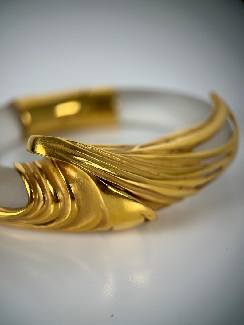 May include: A clear, transparent bangle bracelet with gold-coloured accents. The gold elements feature a textured, layered design. The bracelet is set against a white background, highlighting the contrast between the clear and gold materials.