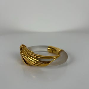 May include: A clear, circular bracelet with a gold-coloured, layered design on one side and a gold clasp. The bracelet has a frosted appearance and is likely made of acrylic or a similar material. The gold elements add a touch of elegance.