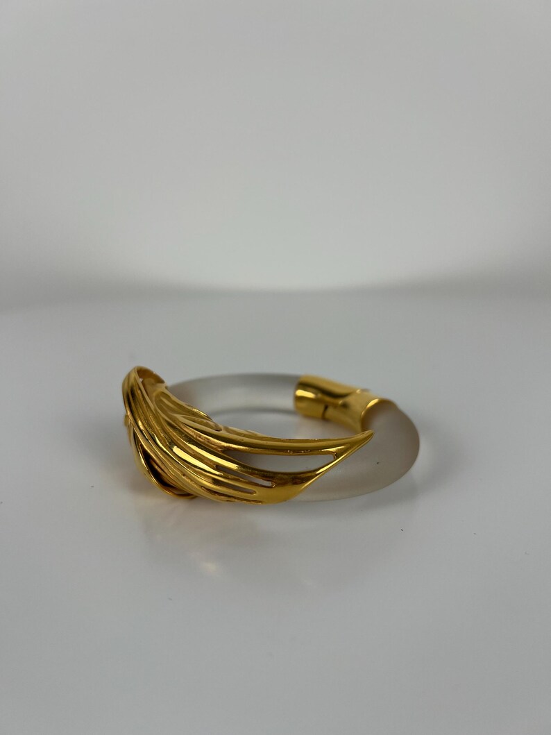 May include: A clear, circular bracelet with a gold-coloured, layered design on one side and a gold clasp. The bracelet has a frosted appearance and is likely made of acrylic or a similar material. The gold elements add a touch of elegance.