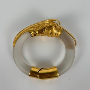 May include: A gold-toned metal and frosted glass bracelet. The bracelet has a circular design with a gold clasp and decorative gold accents. The gold elements have a textured, organic design.
