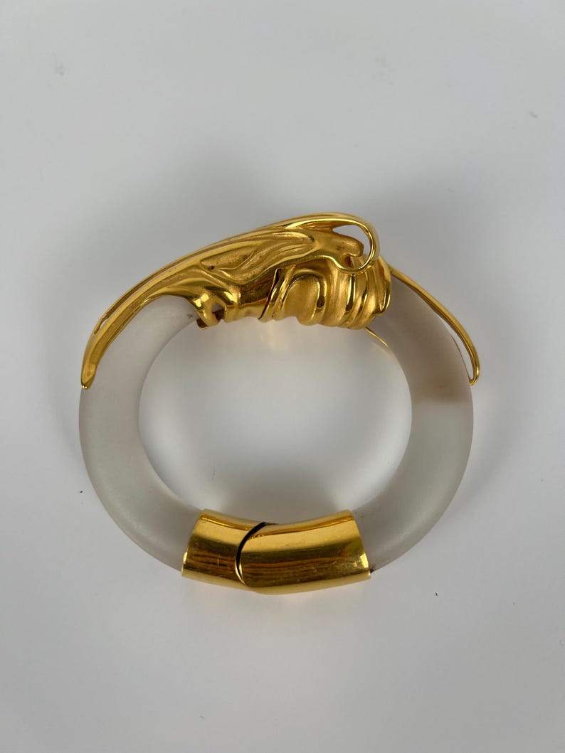 May include: A gold-toned metal and frosted glass bracelet. The bracelet has a circular design with a gold clasp and decorative gold accents. The gold elements have a textured, organic design.