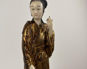 May include: A detailed figurine of a woman in a gold-coloured robe. The figure has a serene expression, with black hair styled in a bun. The robe has a textured appearance, with gold and brown tones. The figurine is standing.
