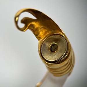 May include: Close-up of a gold ring with a distinctive design. The ring has a rounded, ribbed band and a decorative top with a swirling shape. The ring is set against a white background.