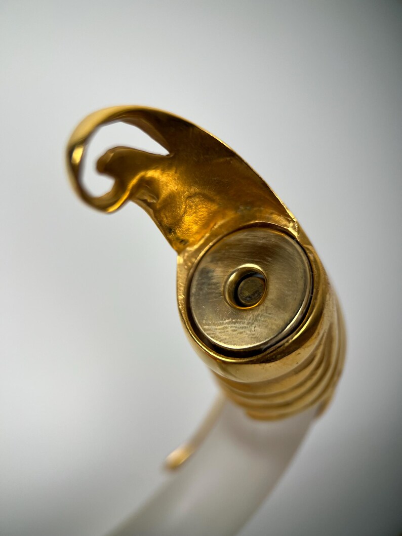 May include: Close-up of a gold ring with a distinctive design. The ring has a rounded, ribbed band and a decorative top with a swirling shape. The ring is set against a white background.