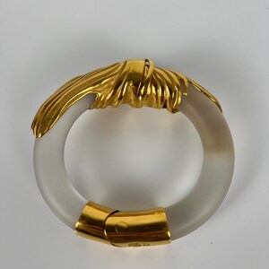 May include: A gold-coloured metal and frosted glass bracelet. The bracelet has a circular design with a gold-coloured knot detail and a gold clasp. The frosted glass has a translucent appearance.