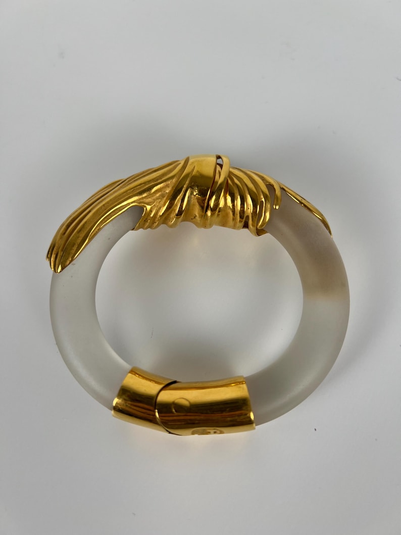 May include: A gold-coloured metal and frosted glass bracelet. The bracelet has a circular design with a gold-coloured knot detail and a gold clasp. The frosted glass has a translucent appearance.