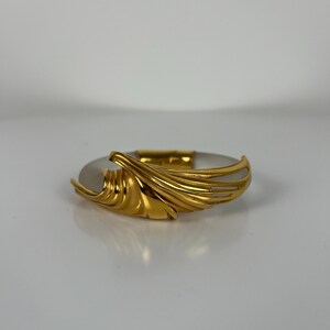May include: A gold-coloured bracelet with a central oval, light-coloured stone. The bracelet features a decorative, flowing gold design on one side, adding an elegant touch. The piece is set against a neutral background, highlighting its design.