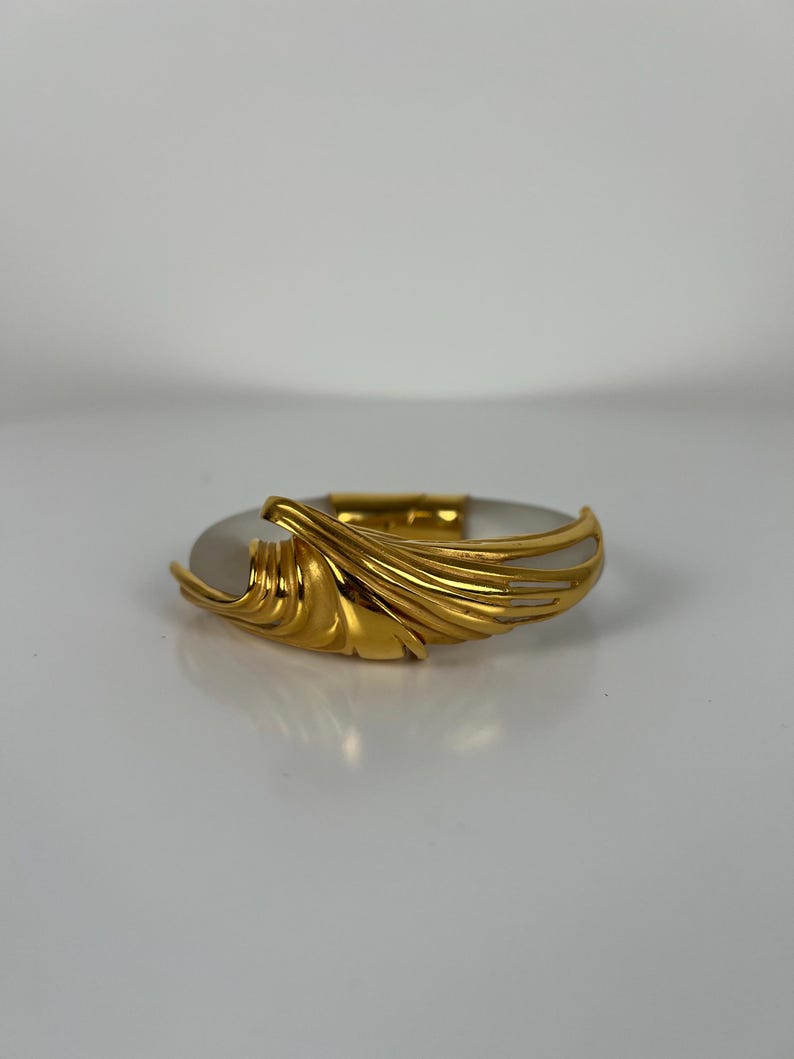 May include: A gold-coloured bracelet with a central oval, light-coloured stone. The bracelet features a decorative, flowing gold design on one side, adding an elegant touch. The piece is set against a neutral background, highlighting its design.