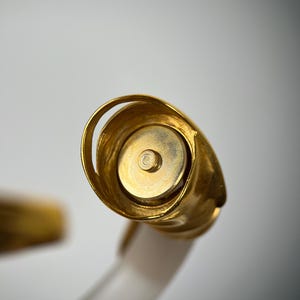 May include: Close-up of a gold bottle stopper with a rounded, spiral design. The stopper has a circular base and a small, central screw. The handle is white and translucent.