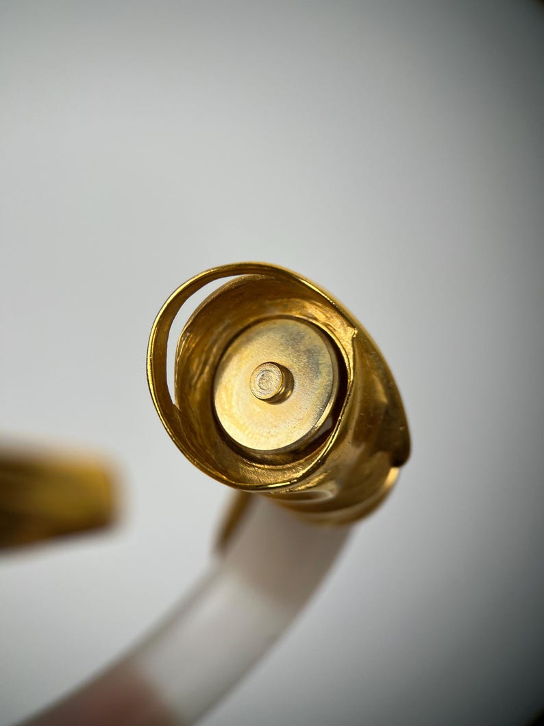 May include: Close-up of a gold bottle stopper with a rounded, spiral design. The stopper has a circular base and a small, central screw. The handle is white and translucent.