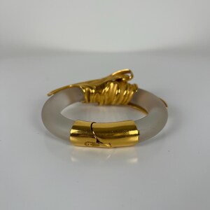 May include: A gold and frosted glass bangle bracelet. The bracelet has a hinged clasp and a decorative gold accent. The gold accents are intricately designed, adding elegance. The frosted glass gives the bracelet a unique, modern look. The bracelet is approximately 6 cm in diameter.