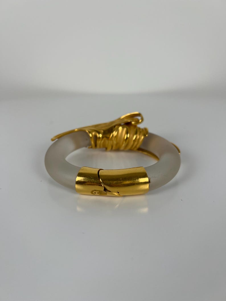 May include: A gold and frosted glass bangle bracelet. The bracelet has a hinged clasp and a decorative gold accent. The gold accents are intricately designed, adding elegance. The frosted glass gives the bracelet a unique, modern look. The bracelet is approximately 6 cm in diameter.