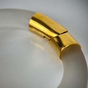 May include: A clear, frosted bangle bracelet with a gold-coloured clasp. The bracelet has a smooth, rounded shape and a subtle, translucent quality. The gold clasp adds a touch of elegance to the simple design. This accessory is suitable for everyday wear.