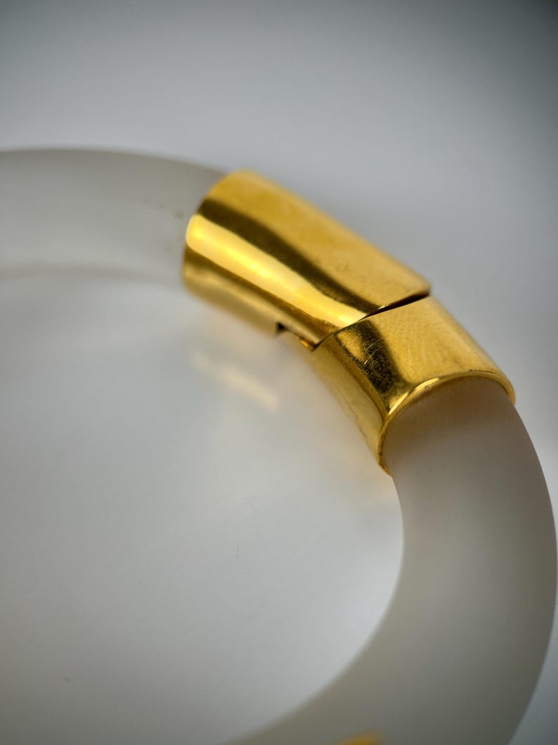 May include: A clear, frosted bangle bracelet with a gold-coloured clasp. The bracelet has a smooth, rounded shape and a subtle, translucent quality. The gold clasp adds a touch of elegance to the simple design. This accessory is suitable for everyday wear.
