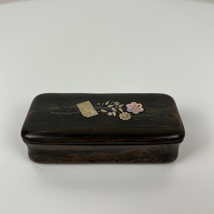 May include: A dark brown rectangular box with a floral design on the lid. The box features a small tag and delicate flowers in pink, white, and gold. The box is likely used for storing small items.