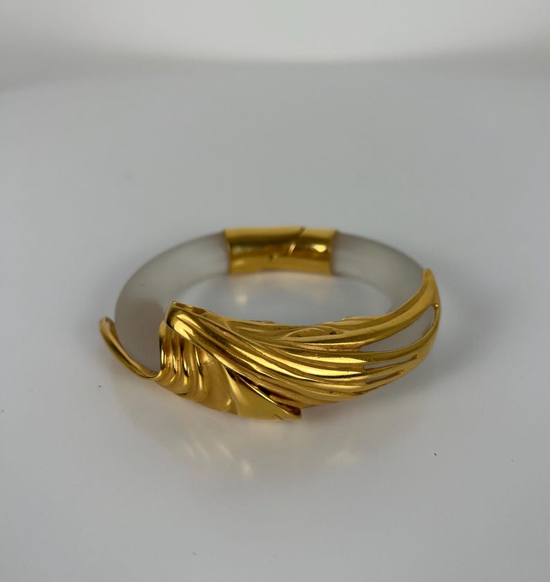 May include: A gold-coloured bracelet with a clear, circular band. The bracelet features a decorative gold leaf design on the front. The clasp and accents are also gold. The jewellery is set against a white background.