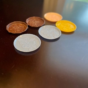 May include: A collection of six decorative coasters in various colors. The coasters are round and feature embossed floral or patterned designs. Colors include brown, orange, yellow, and white. The coasters are arranged on a dark brown surface.