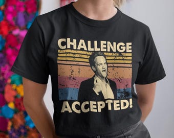 Puede incluir: Camiseta negra con un gráfico de estilo vintage que presenta a un hombre con una expresión de sorpresa. El gráfico dice "Challenge Accepted!" en texto blanco.