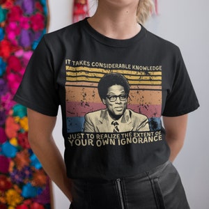 May include: Black t-shirt with a retro design. It features a portrait of a man in glasses and a suit, and the text "IT TAKES CONSIDERABLE KNOWLEDGE JUST TO REALIZE THE EXTENT OF YOUR OWN IGNORANCE" in a vintage style.
