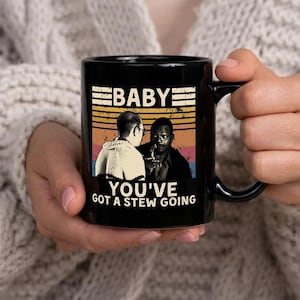 Puede incluir: Taza de cerámica negra con el texto "BABY YOU'VE GOT A STEW GOING" y una imagen de dos hombres. La taza tiene un asa negra y un interior negro. El diseño tiene un fondo retro de puesta de sol.