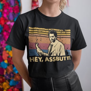 Könnte beinhalten: Schwarzes T-Shirt mit einem Vintage-Grafikdruck eines Mannes, der einen Daumen hoch zeigt, und dem Text "HEY, ASSBUTT!"