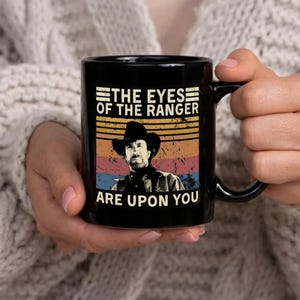 May include: Black ceramic mug featuring a retro design with the text "THE EYES OF THE RANGER ARE UPON YOU." The design includes a portrait of a man in a cowboy hat set against a striped background in shades of orange, yellow, blue, and red.