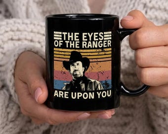Może przedstawiać: Czarny ceramiczny kubek z napisem "THE EYES OF THE RANGER ARE UPON YOU" i portretem mężczyzny w kapeluszu kowbojskim. Projekt ma styl retro z paskowanym tłem w kolorach pomarańczowym, żółtym, niebieskim i czerwonym.