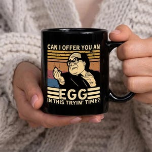 Może przedstawiać: Czarny ceramiczny kubek z obrazem w stylu retro przedstawiającym mężczyznę trzymającego jajko. Kubek ma napis "CAN I OFFER YOU AN EGG IN THIS TRYIN' TIME?" w kolorze białym i żółtym. Kubek jest trzymany przez osobę.