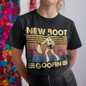May include: Black t-shirt with a vintage-style design. The graphic reads "NEW BOOT GOOFIN" and depicts a police officer with a bag, set against a striped backdrop in yellow, orange, blue, and brown. A humorous and eye-catching design.