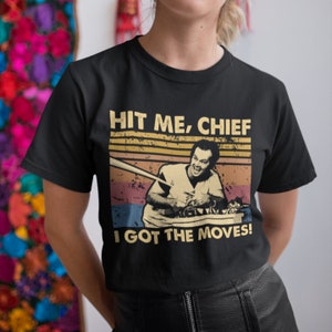 Puede incluir: Camiseta negra con un gráfico de estilo vintage de un hombre sosteniendo un bate de béisbol. El gráfico dice "Hit Me, Chief I Got The Moves!"
