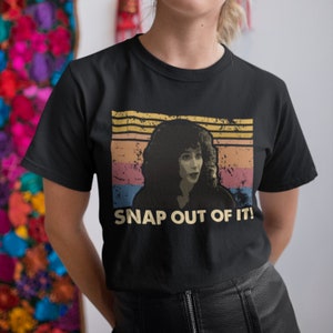 以下が含まれることがあります： 「SNAP OUT OF IT!」と書かれた、カールした髪の女性が描かれたヴィンテージスタイルのグラフィックがプリントされた黒いTシャツ