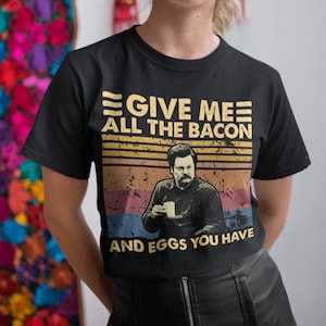 Può includere: Una maglietta nera con una grafica in stile vintage che presenta un uomo con i baffi e il testo "Give me all the bacon and eggs you have."