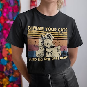 May include: Black t-shirt featuring a retro design with the text "GIMME YOUR CATS AND NO ONE GETS HURT." The graphic includes a cartoon alien character with a threatening pose, set against a striped background.
