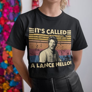 May include: Black t-shirt with a retro design. The text "IT'S CALLED A LANCE HELLO!" is above a vintage image of a man. The design incorporates horizontal stripes in yellow, orange, pink, and blue, creating a vintage aesthetic.