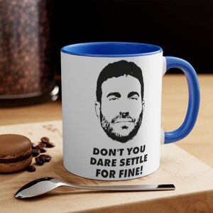 May include: White ceramic mug with a blue rim and handle. The mug features a black and white silhouette of a man's face with the text "Don't you dare settle for fine!"
