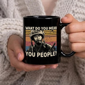 May include: Black ceramic mug featuring a retro-style graphic of a soldier with the text "WHAT DO YOU MEAN YOU PEOPLE?". The design includes a sunset-like background with orange, yellow, and blue stripes. The mug has a black handle.