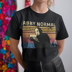 May include: Black t-shirt with a vintage-style graphic of a person holding a glass of liquid. The graphic has the text "Abby Normal" in white letters.
